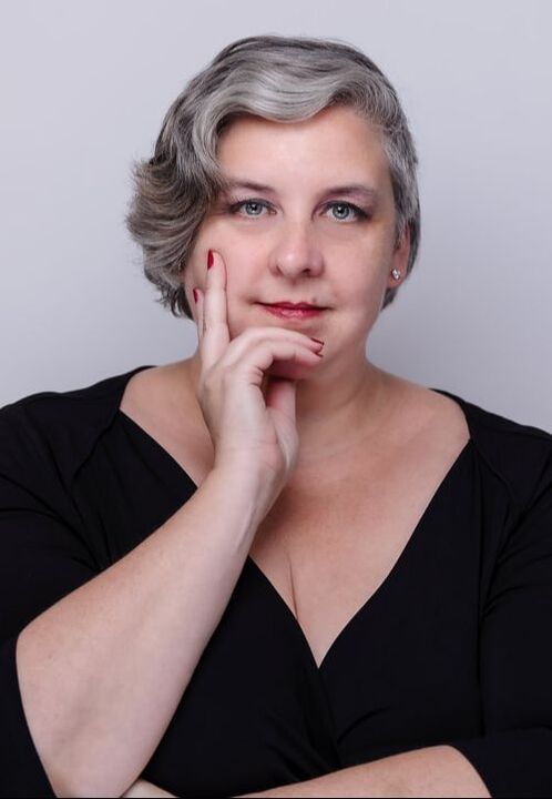 Photo of Jessica Ellis-Wilson, a pluz-sized white nonbinary person with blue eyes and short grey hair swept to the left, looking directly at the camera and resting their chin on their right hand while their left arm rests across their body with their left hand under their right elbow.  They are standing in front of a grey-white background in a black wrap shirt with 3/4 sleeves. They have red nail polish on short nails, matching red lipstick, and a single stud earring is visible in their left ear.