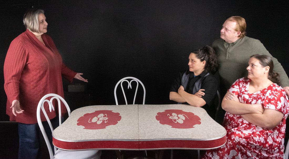 Photo of a black box space with a white formica table with red detailing on the sides and two red and white floral details in the center of each side of the table. There are four white metal “ice cream shop” chairs around the sides and upstage edge of the table. There are four people around the table. From left to right, figure one is Jess Wilson (playing Ruth) is standing, facing the other figures with their arms open to either side. Jess is a plus-sized nonbinary person, wearing dark blue jeans and a dark red sweater, and their hair is silver cut in a chin-length bob. The remaining three figures are looking at Jess. Figure two is Victoria Smith (playing Beverly), seated with arms crossed over her chest, wearing a black and grey bowling shirt over a teal undershirt. She has long, curly, black hair which is pulled back into a pony tail. Figure three is Garrett Olson (playing Jimmy), standing behind Victoria and Stephanie with a hand resting on either chair. He has dark auburn hair, cut short, and is wearing a sage green henley. Figure four is Stephanie Clark (playing Clara), seated at the end of the table with her arms crossed over her chest. She is wearing a short-sleeved red housedress with a large floral pattern in white, and has long, dark brown hair which is pulled back in a pony tail.