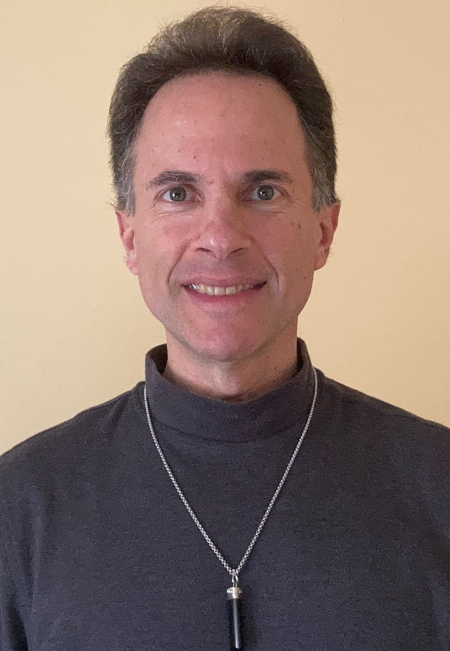 Photo of Ian Rubinstein, a white man with hazel eyes and short brown hair swept back from the face, looking directly at the camera with a small smile. His head, shoulders, upper torso, and upper arms are visible against a solid dark yellow background. He is wearing a dark grey mock turtleneck with a black crystal rod with silver fitting and silver chain.